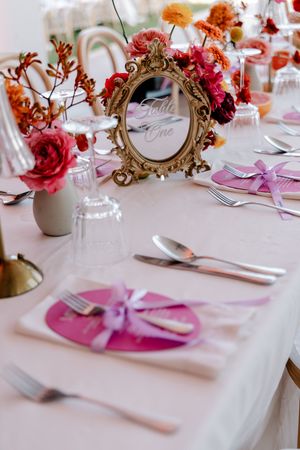 Elegant wedding table setting with pink plates, gold ornate table number mirror, and colorful floral centerpieces.