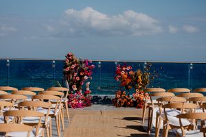 Outdoor beachfront wedding ceremony setup with colorful floral arch, wooden chairs, and ocean backdrop under blue sky.