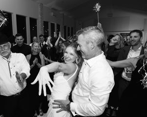 Black and white photo of a bride and groom dancing joyfully at their wedding reception surrounded by celebrating guests.