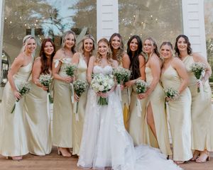 Bride in white lace gown poses with eight bridesmaids in matching champagne satin dresses holding white floral bouquets.