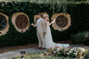 A couple in wedding attire share an intimate moment against a garden wall with circular wooden accents and floral decor.