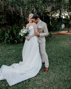 A couple in wedding attire share a romantic moment outdoors on a grassy lawn surrounded by trees.