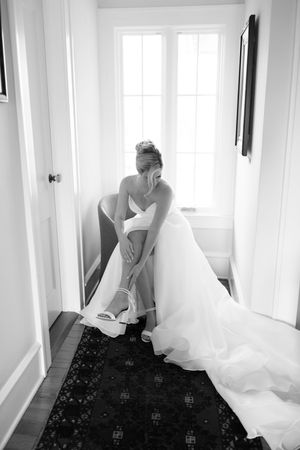 Black and white photo of a person adjusting a flowing white wedding dress in a hallway with dark patterned carpet.