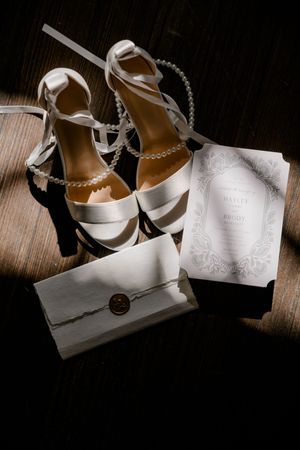 Elegant white bridal shoes and wedding invitation cards arranged artistically on dark wooden surface with dramatic lighting.