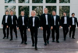 Groom and groomsmen in black tuxedos with bow ties walking confidently on a wooden deck at a wedding venue.