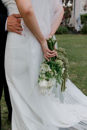 Close-up of bride in white wedding dress holding elegant white floral bouquet while groom embraces her from behind.