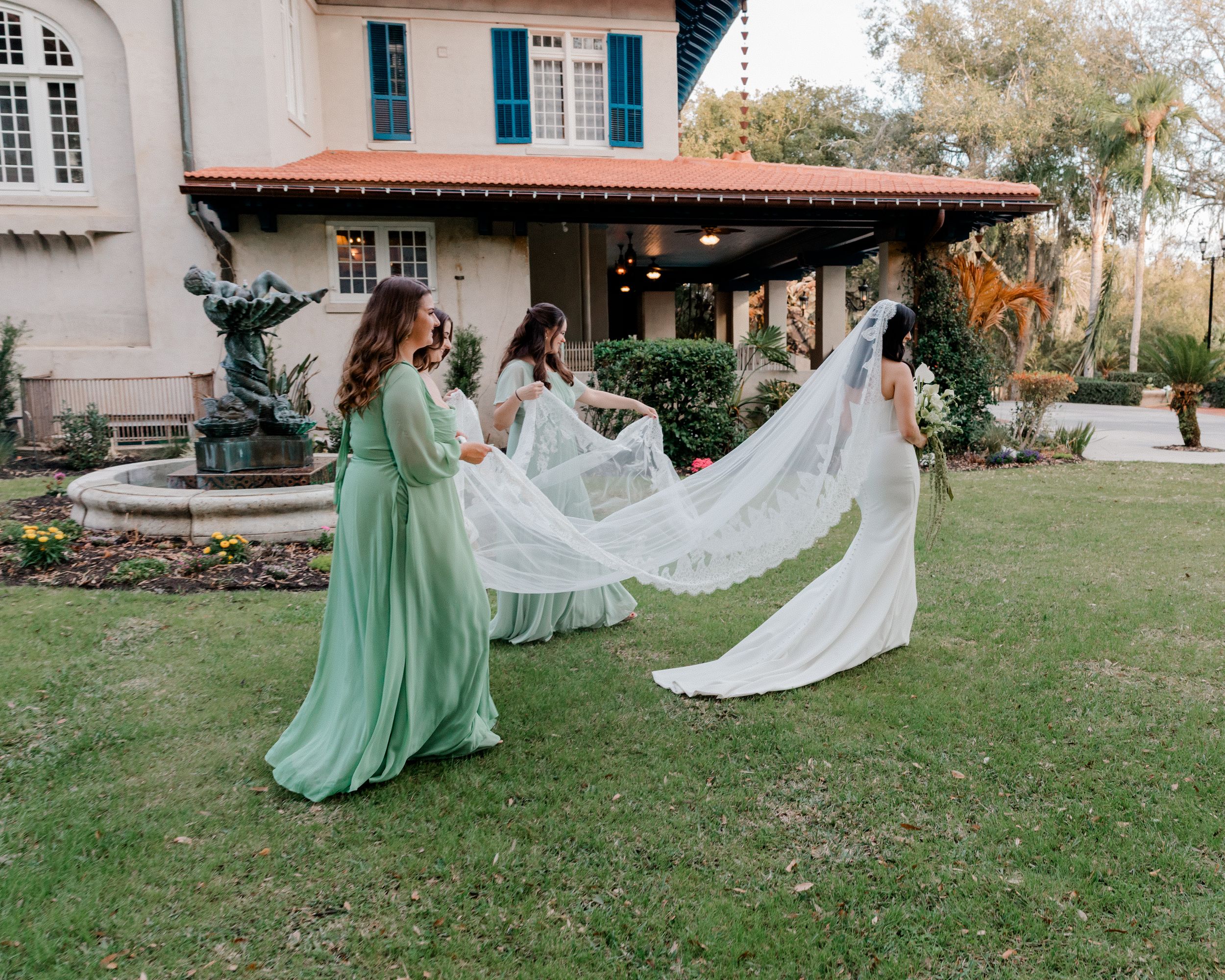 Bridesmaids in sage green dresses hold a long lace bridal veil outdoors near a fountain at a wedding venue.