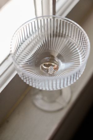 A diamond ring sits in a crystal glass with radial ribbed pattern on a white windowsill.