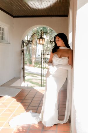 Bride in elegant white strapless gown with gloves poses by arched doorway in sunlit Spanish-style venue.