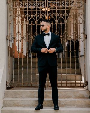 Stylish man in black tuxedo with bow tie posing before ornate iron gates on stone steps.