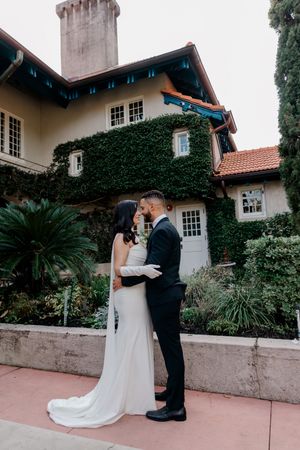 A romantic wedding couple embraces in front of an ivy-covered mansion with Spanish-style architecture and terra cotta patio.