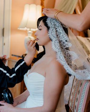 Bride in strapless gown gets lipstick applied while veil is placed on her dark hair before wedding.