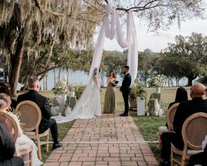 Outdoor wedding ceremony setup with vintage chairs and flowing white fabric draped between trees along a brick pathway.