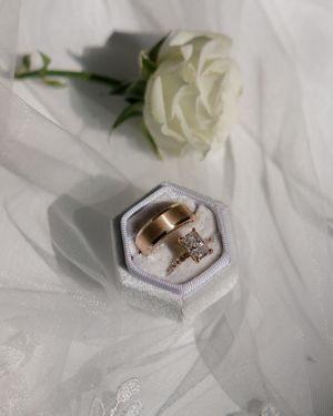 A white rose and wedding rings displayed on delicate hexagonal ring boxes against a light fabric background.