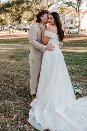 Couple in wedding attire embrace outdoors on grassy lawn during golden hour with trees in background.