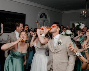 Bride and groom drinking shots with wedding guests in tuxedo and white dress at indoor reception celebration.