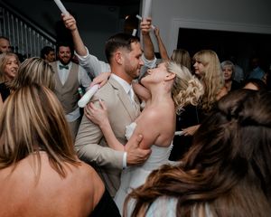 Bride and groom share a romantic kiss surrounded by cheering wedding guests on the dance floor.