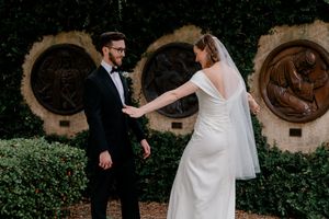 A romantic wedding couple dances together in a garden setting with stone archways and lush greenery in the background.