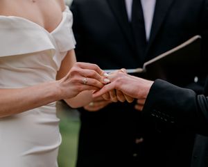 Close up of hands exchanging wedding rings during outdoor marriage ceremony with bride in white and groom in black suit.