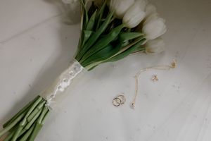 White tulip bouquet with wedding rings and delicate jewelry on a light marble surface.