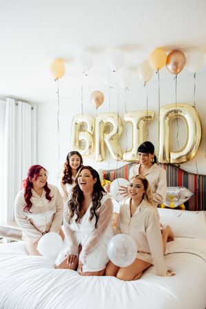 A group in white robes laughs together on a bed with gold balloon letters decorating the wall behind them.