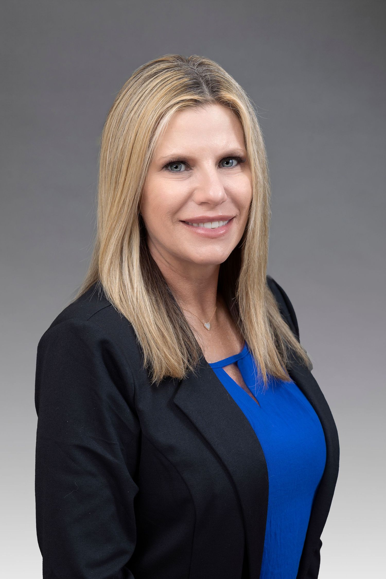 Professional headshot of a person in a black blazer and blue top against a gray background.