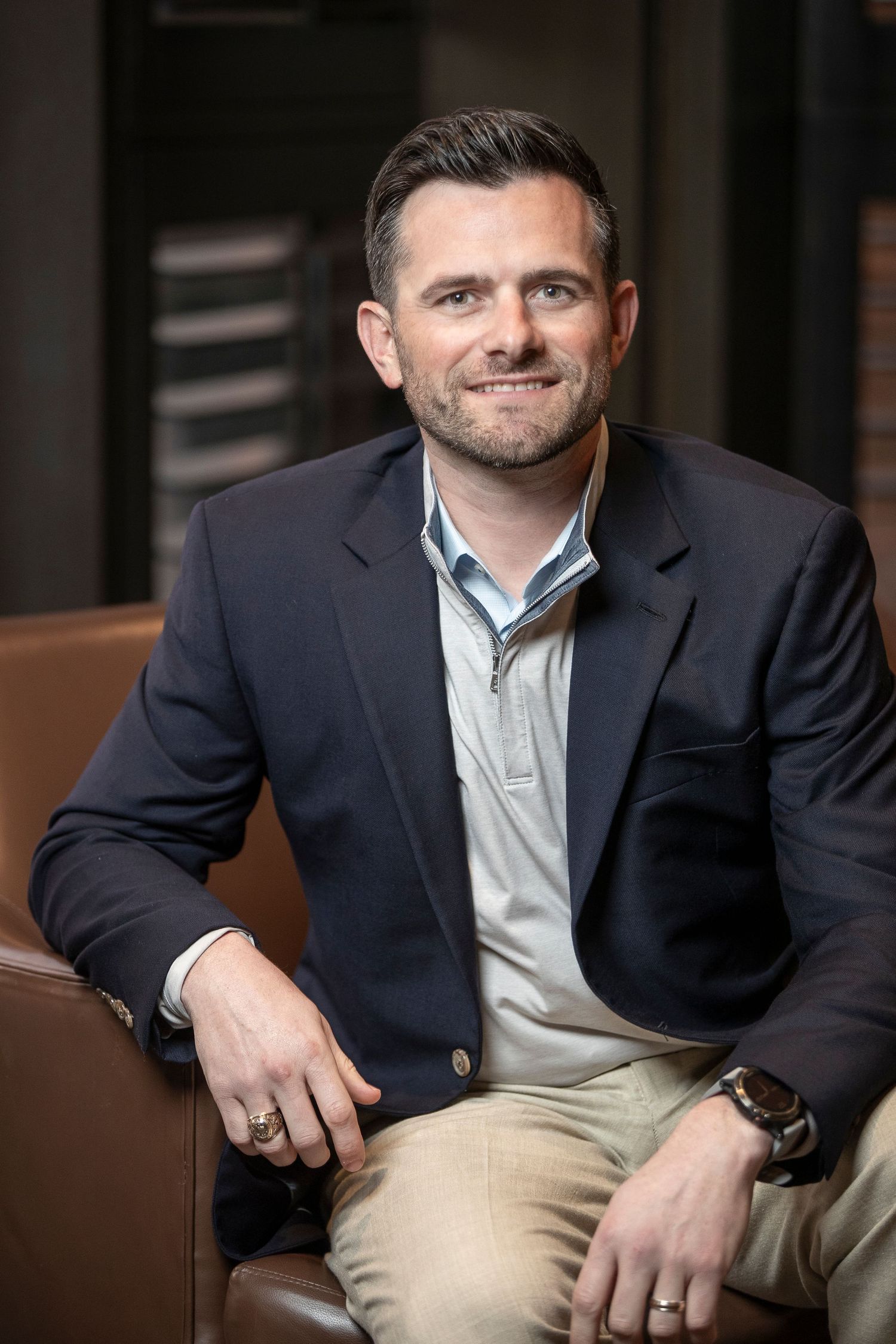 Professional headshot of someone in a navy blazer and light shirt against a dark background.
