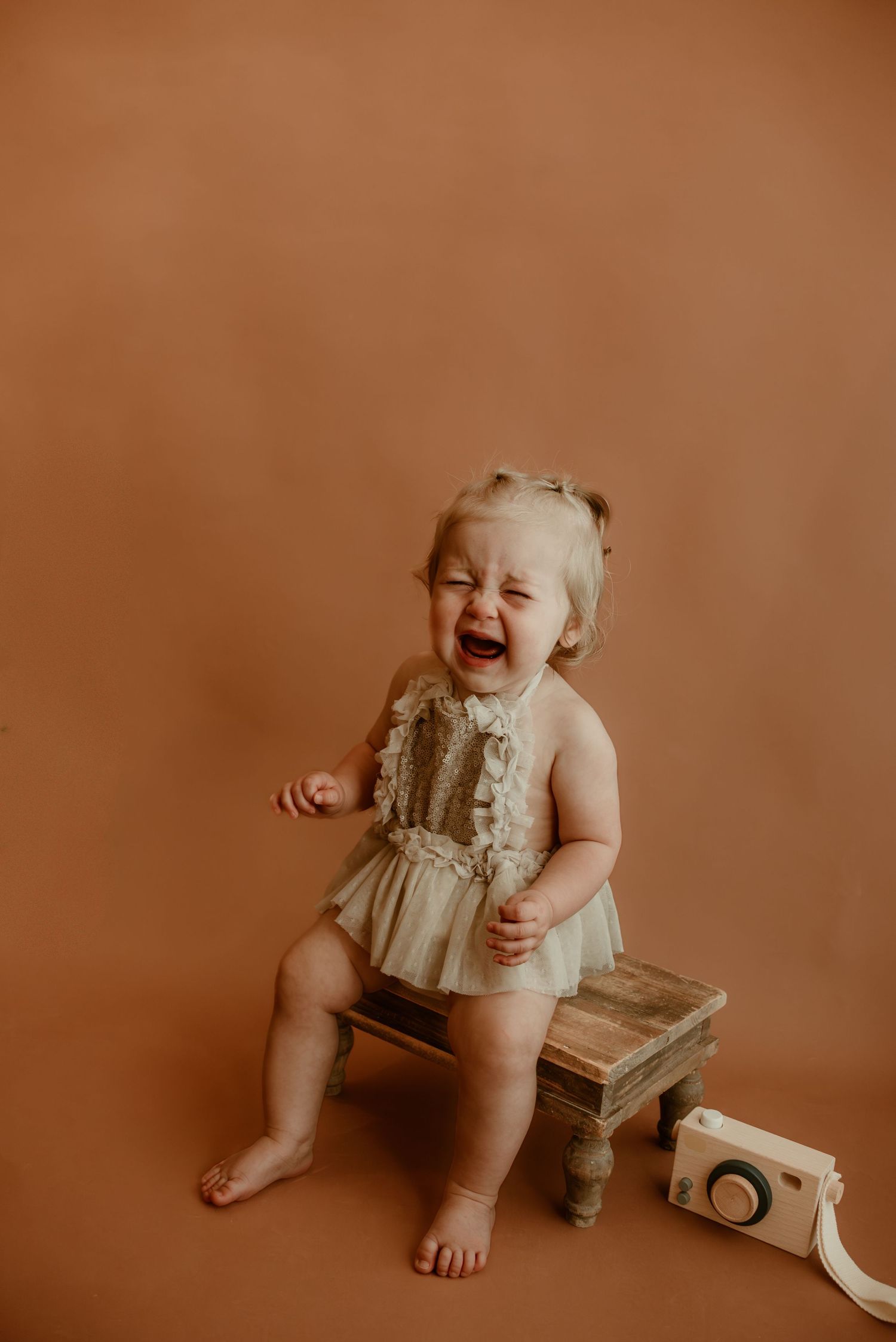Laughing toddler in vintage-style outfit sitting on wooden stool against brown backdrop.