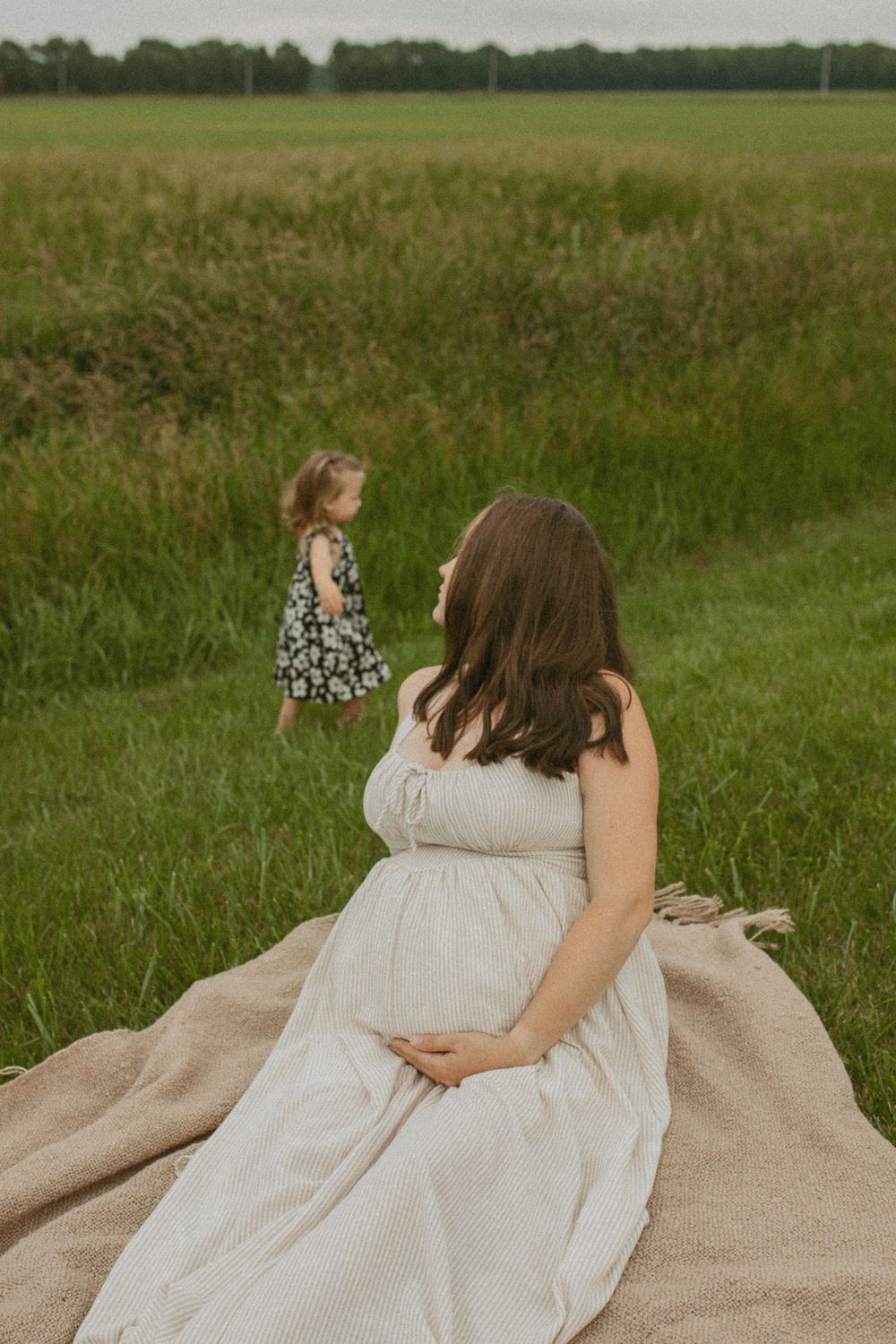 A pregnant woman in a white dress sits on a blanket in a green field watching a toddler play.