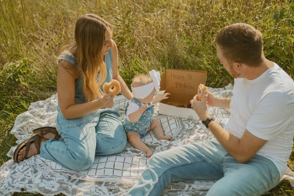 Young family enjoying a picnic in a meadow, sharing donuts with their baby on a lace blanket.