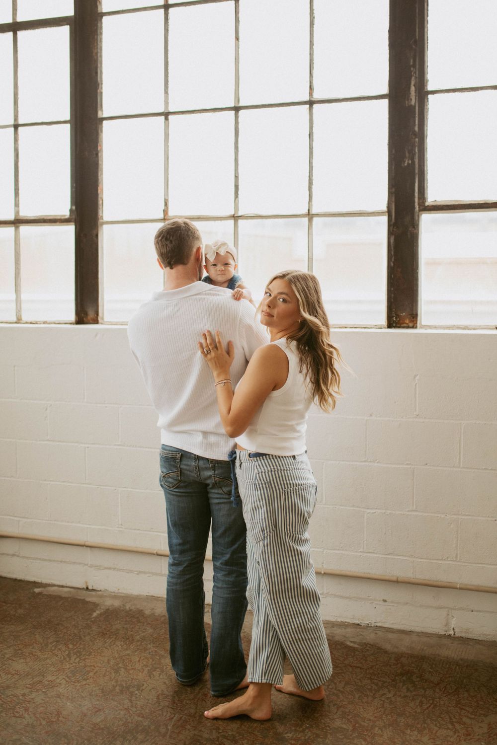 Young family in matching white tops standing by large industrial windows, mother looking back at camera.