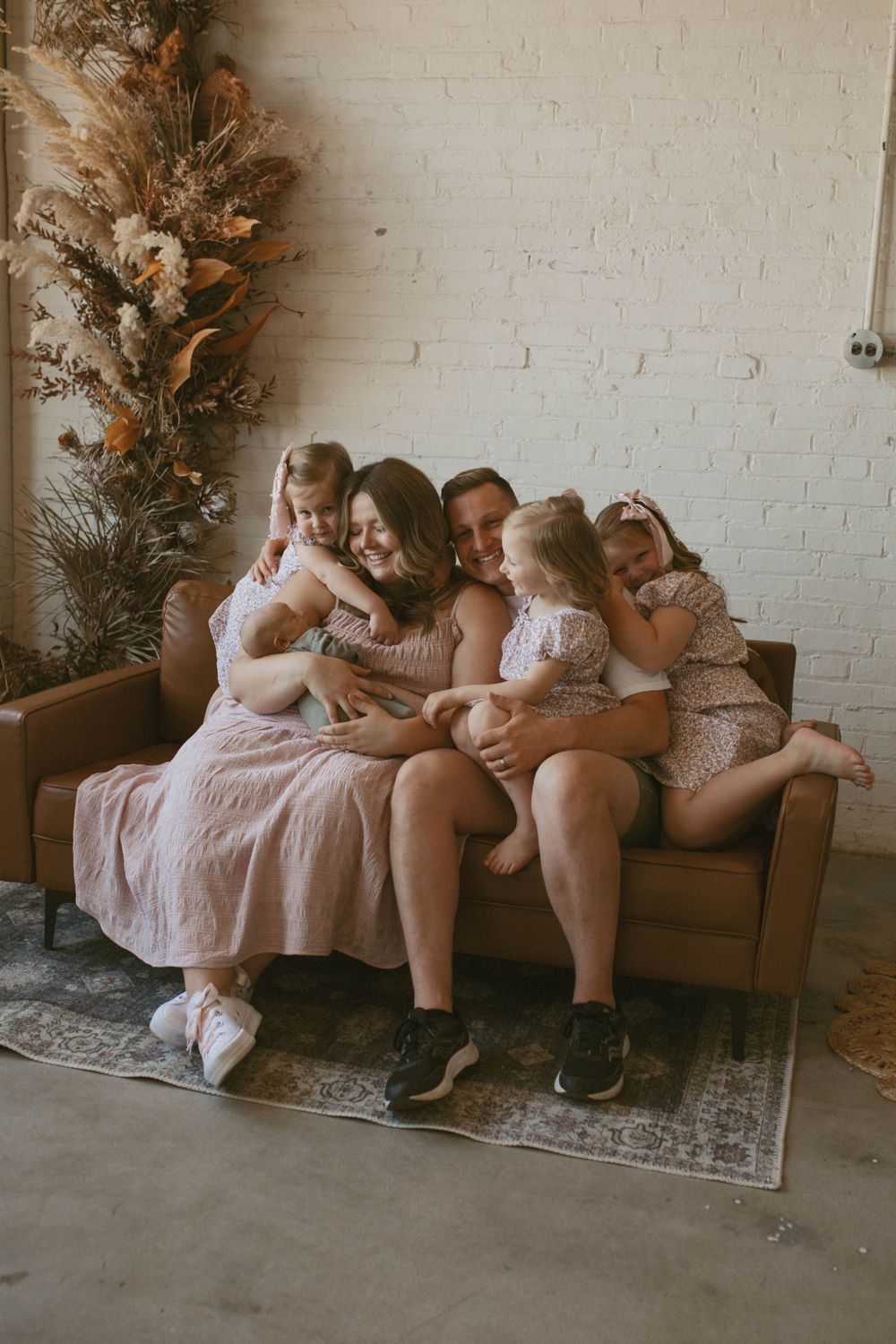 Happy family of five cuddling together on a brown leather sofa with boho dried floral decor.