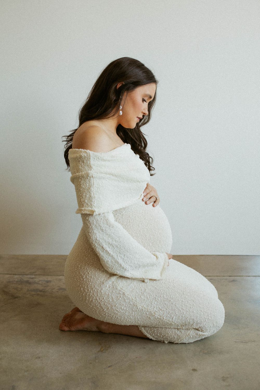Pregnant woman in cream off-shoulder knit dress kneeling on floor cradling baby bump in serene maternity photo.