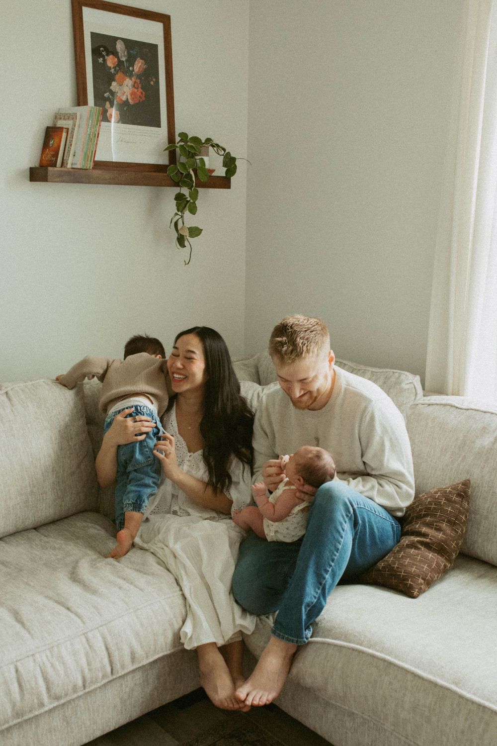 A happy family with a toddler and newborn baby sitting together on a cozy white sofa at home.