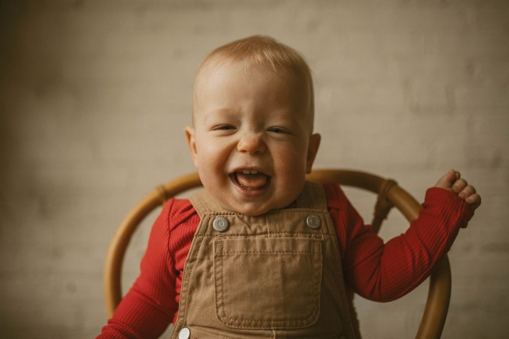 Adorable laughing baby in brown overalls and red shirt sitting on a wooden chair against a white background.