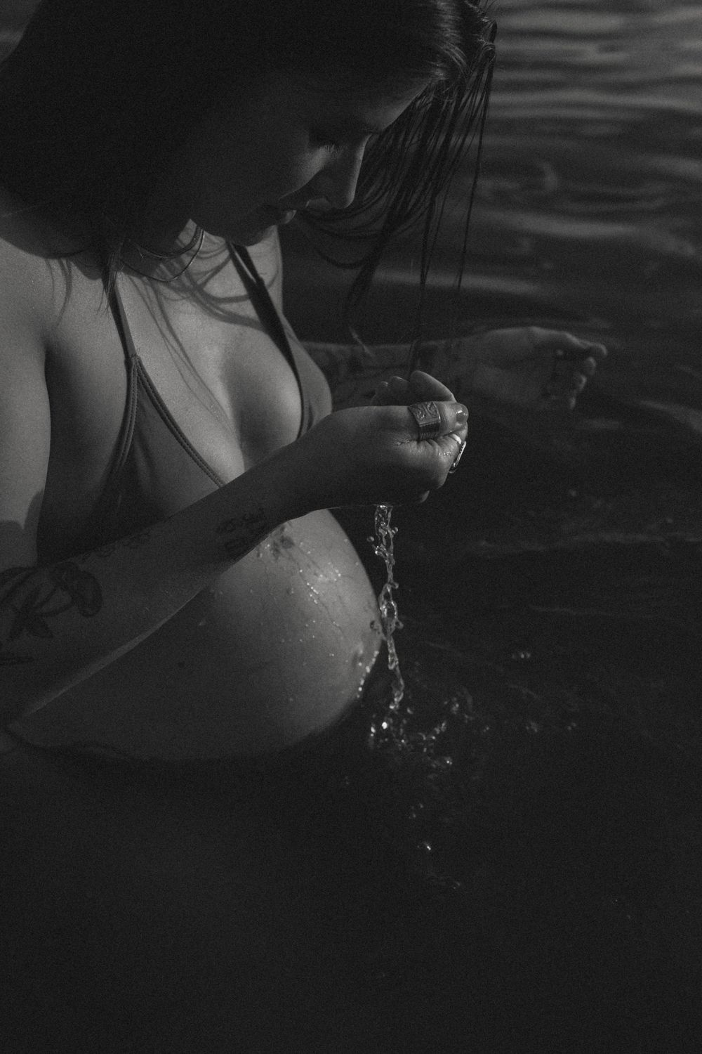A black and white photo of a pregnant woman in a bikini top splashing water in a lake.