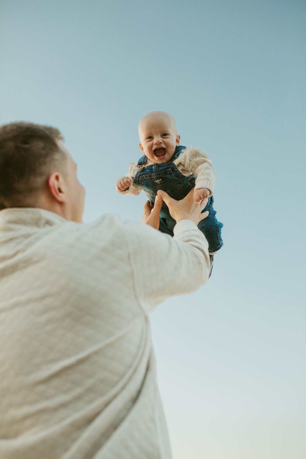 Joyful father lifting laughing baby in denim overalls against a clear blue sky.