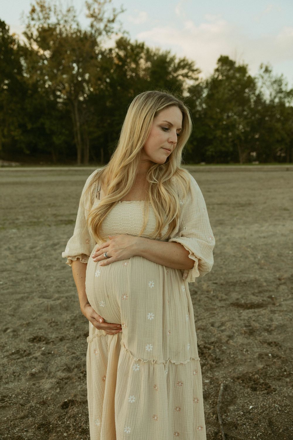 Pregnant woman in cream floral dress cradling baby bump in an open field.