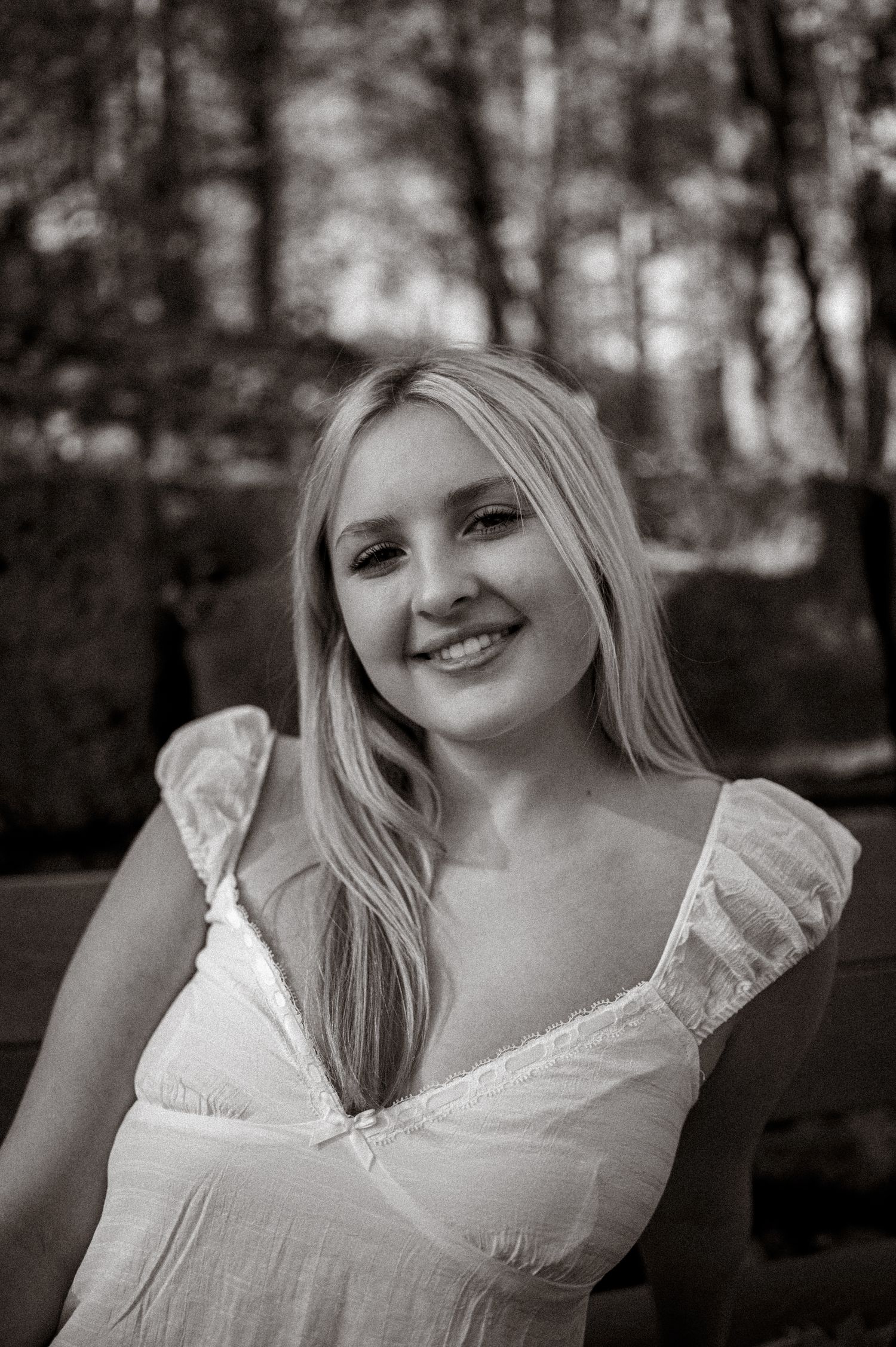 Smiling blonde woman in white ruffled dress photographed outdoors among trees, elegant black and white portrait photography.