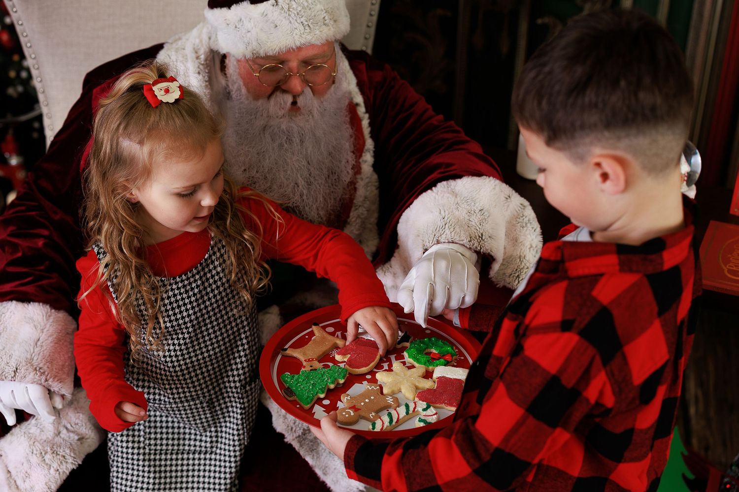 Children in red plaid holiday outfits gather around a Christmas tree opening presents during a festive celebration.