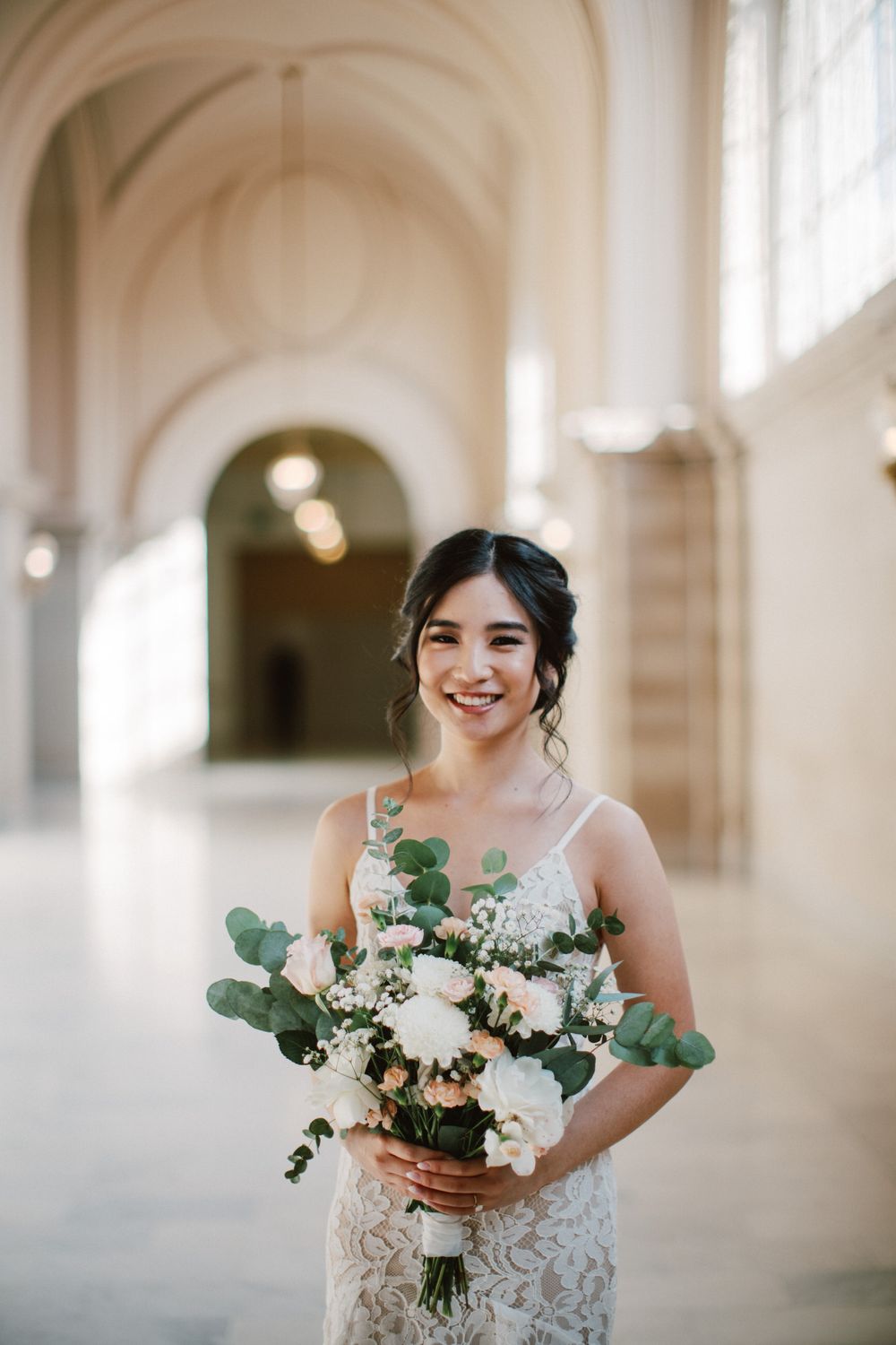 A bride in a lace wedding dress holds a beautiful bouquet featuring white flowers and eucalyptus in an elegant hallway.