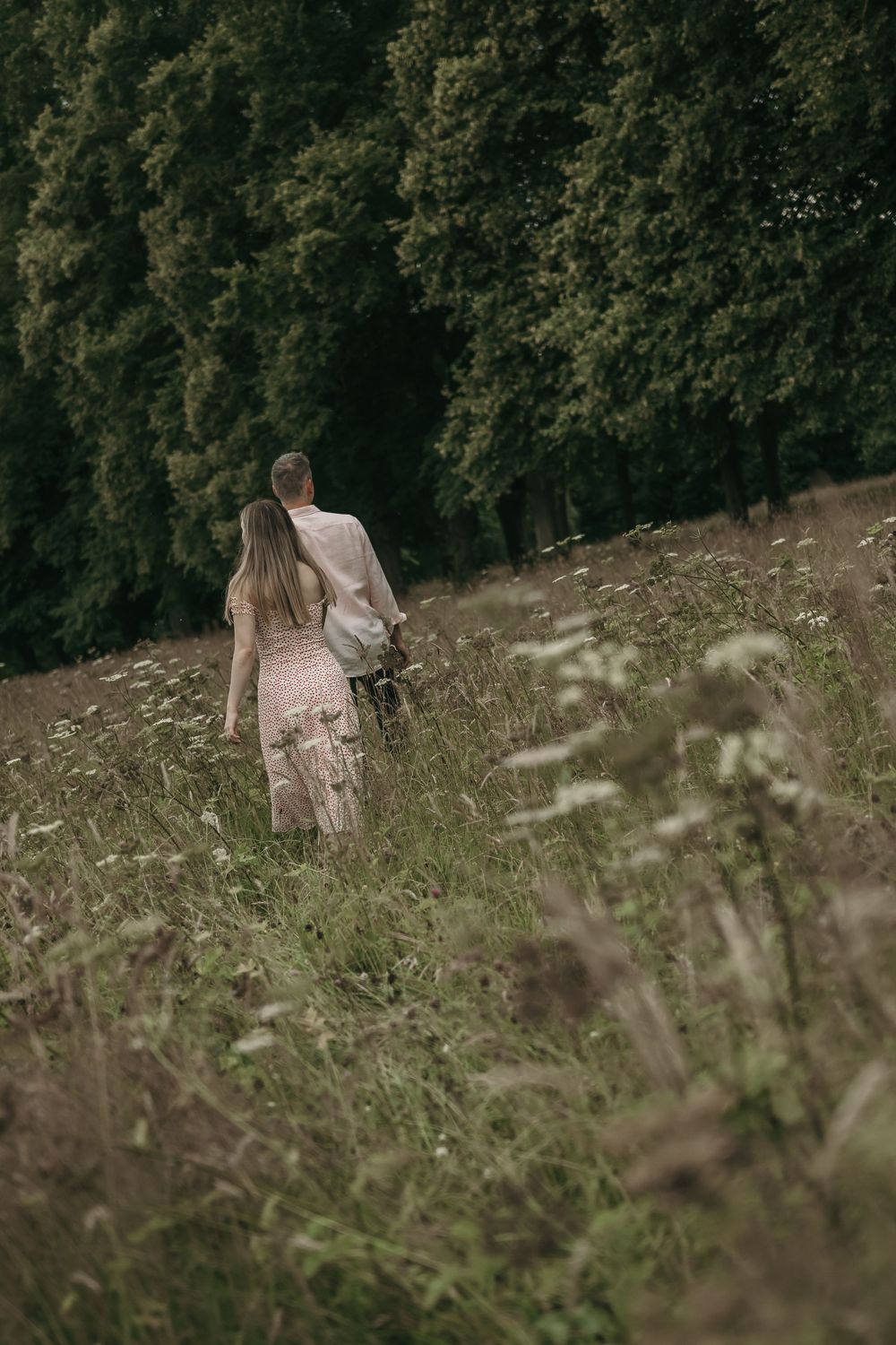 A person in a white dress walks through a meadow with tall grass and trees in the background.