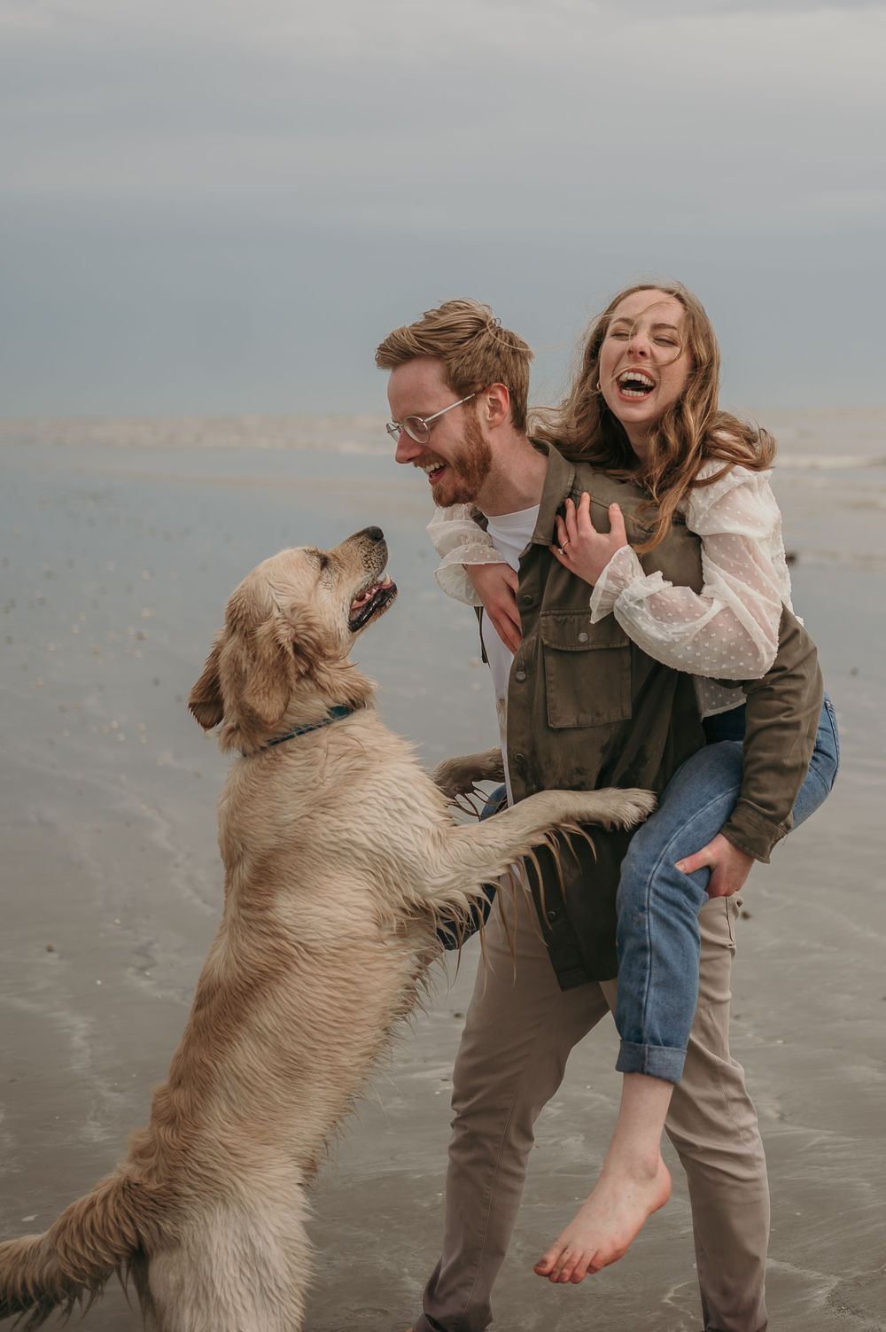 A joyful couple plays with their energetic golden retriever on a misty beach, laughing as the dog jumps up playfully.