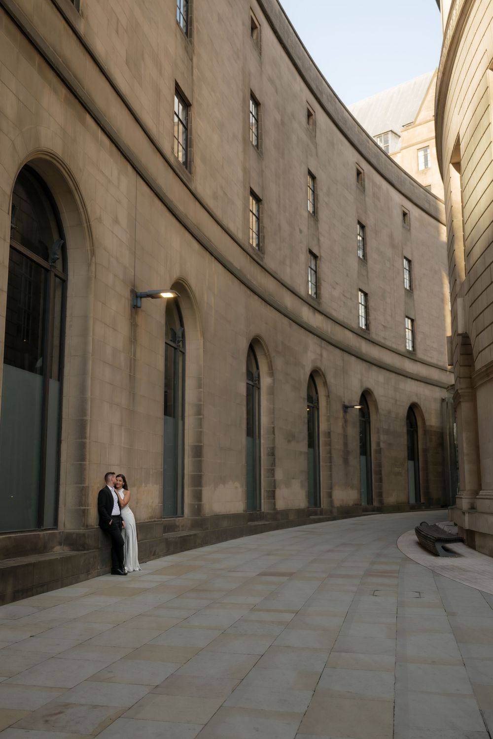 A couple stands against a curved stone wall with arched windows in an elegant architectural setting.