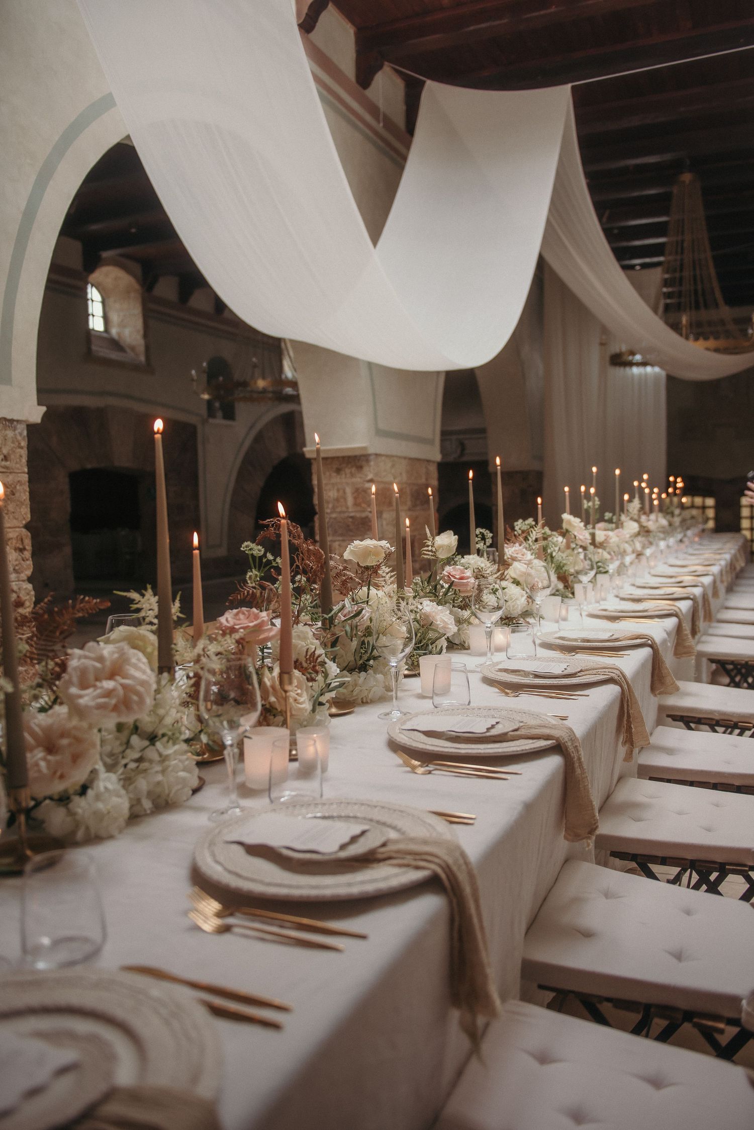 Elegant wedding reception table setting with white draping, candlesticks and floral centerpieces in a rustic stone venue.