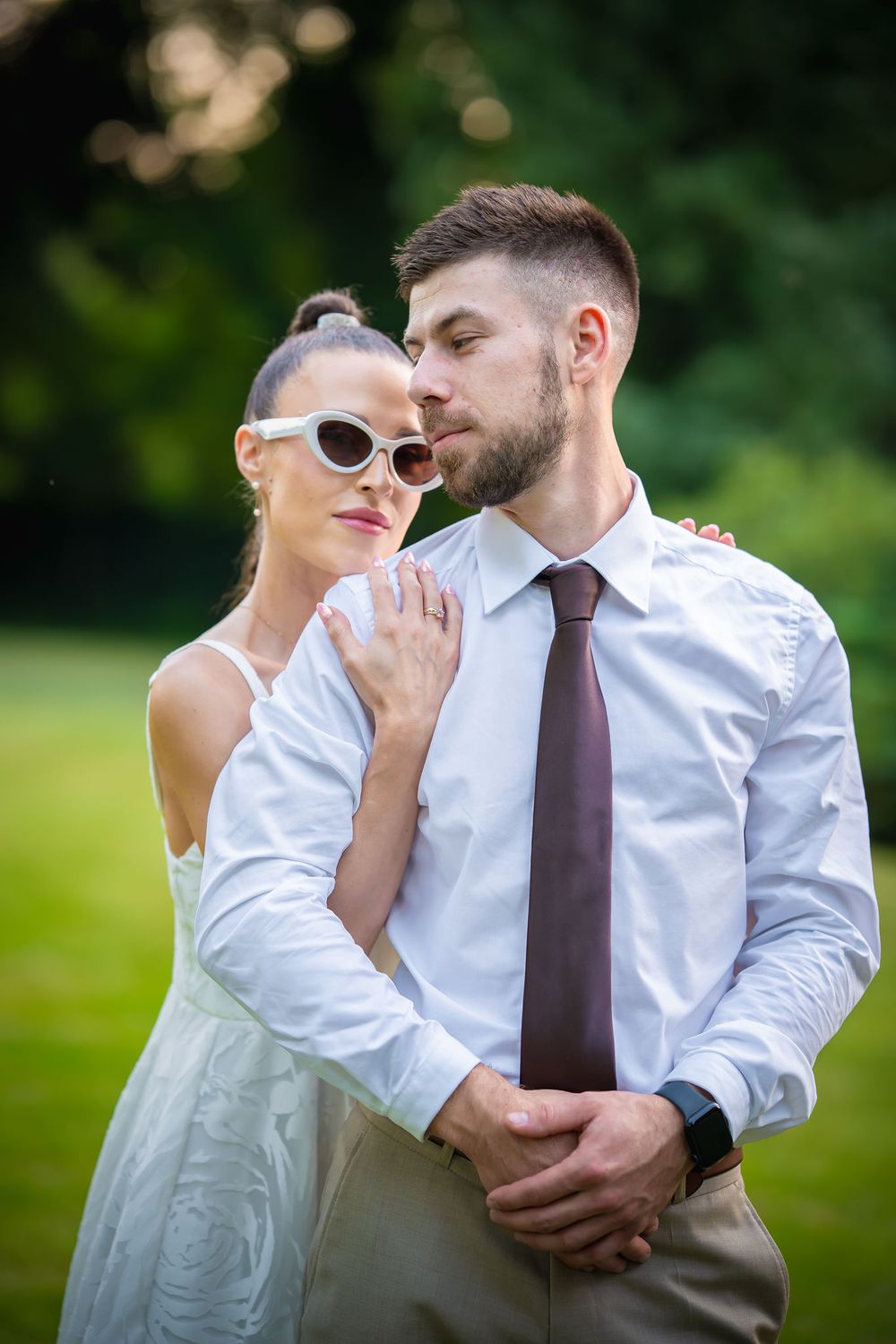 A stylish couple poses outdoors in white attire, with sunglasses and a purple tie against a green natural background.