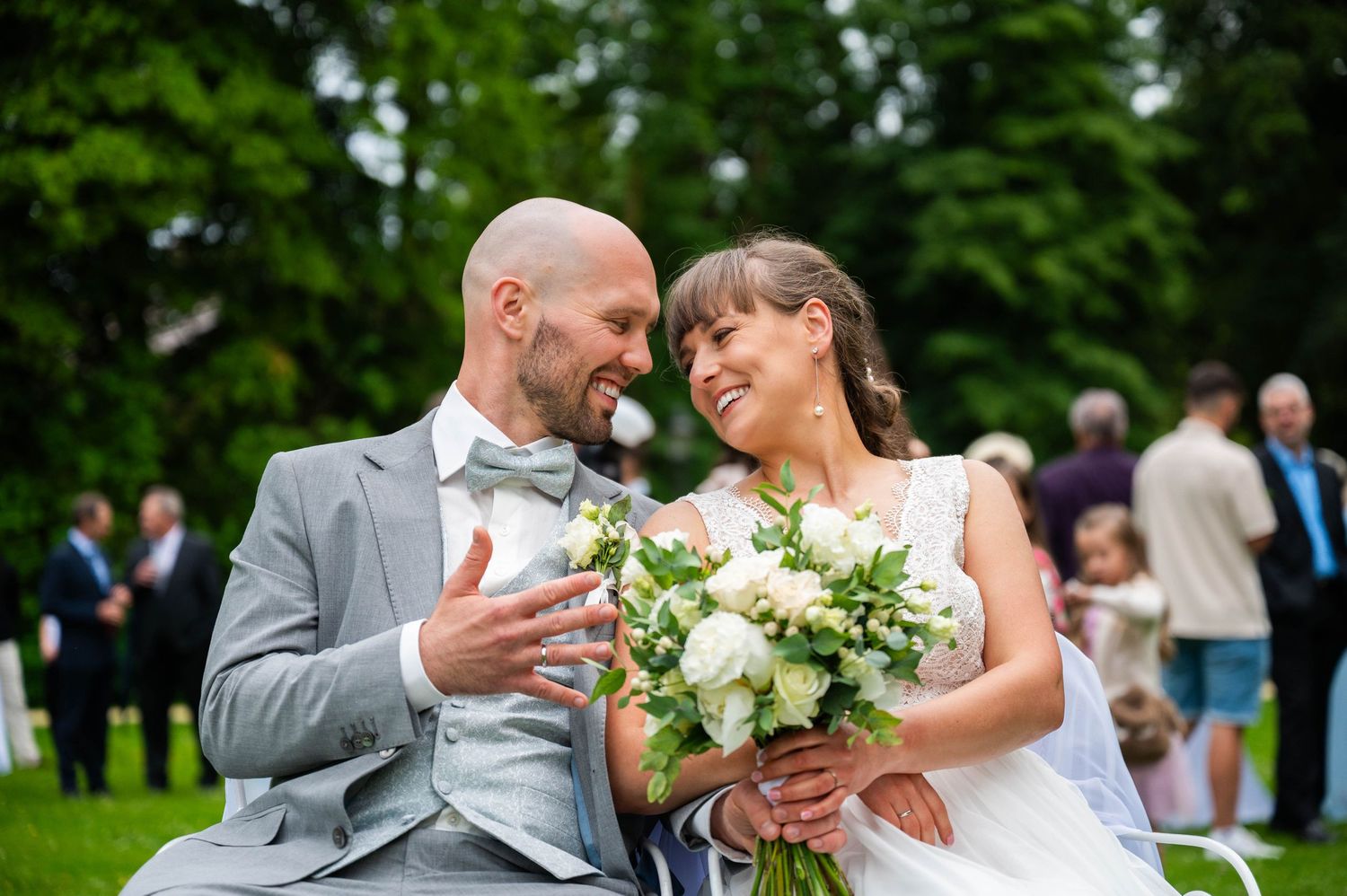 Bride and groom sharing a joyful moment while holding white floral bouquet during outdoor garden wedding celebration.