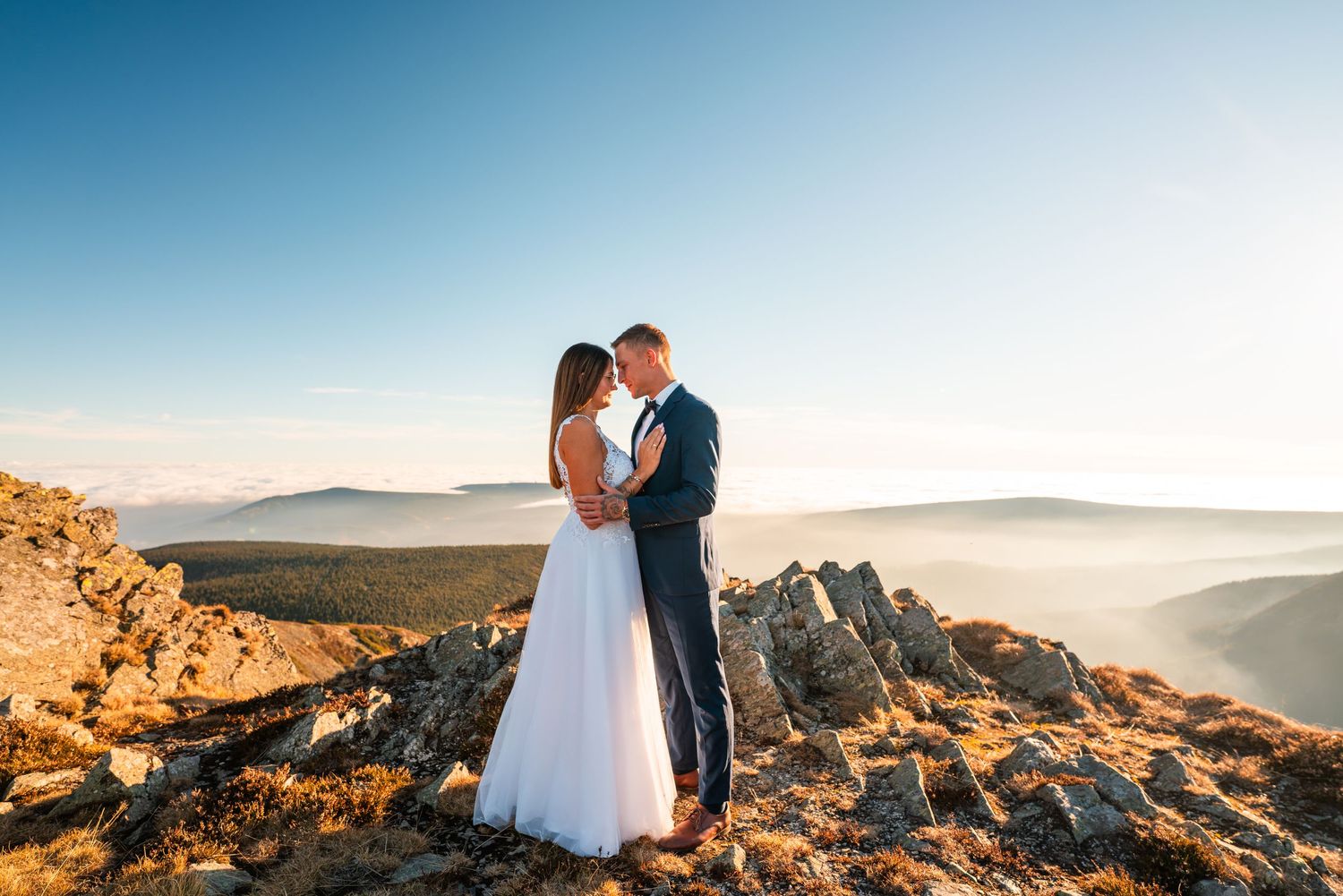 Newlyweds embrace on rocky mountain peak with misty valleys in background.