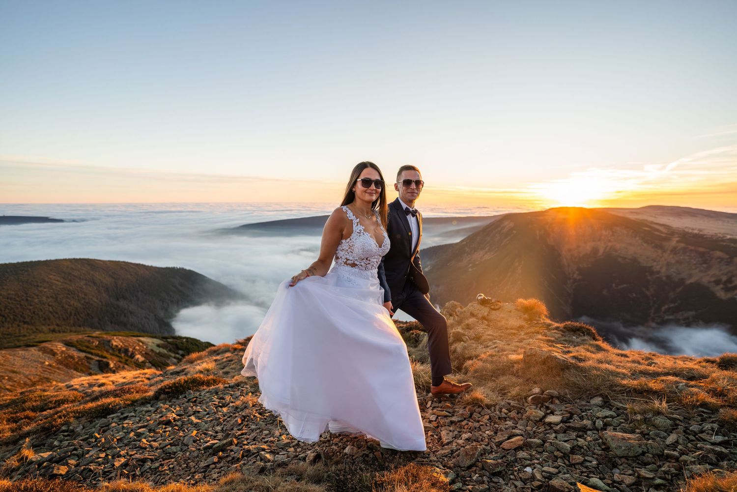 Wedding couple stands on mountain summit with fog-filled valleys below at sunrise.