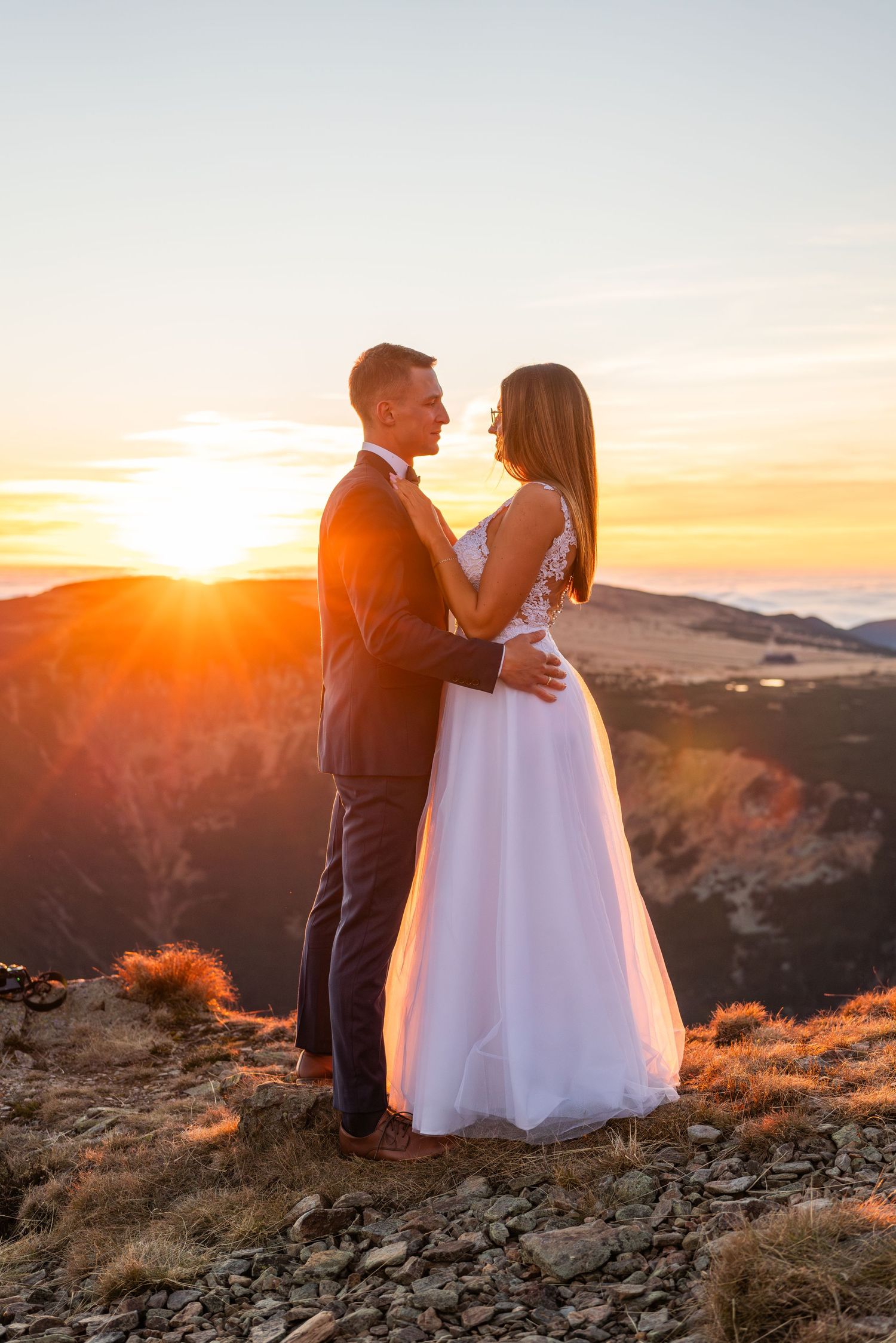 Couple in wedding attire embraces on mountain peak during golden sunset.